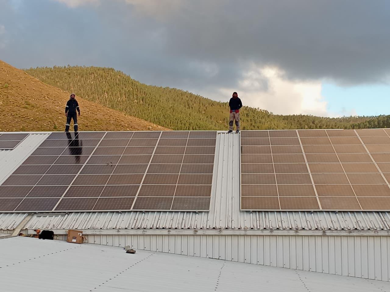 Two team members beside solar panel arrays on a metal roof beneath cloudy skies
